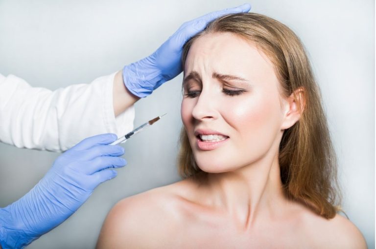 Woman wincing as a clinician wearing gloves approaches her forehead with a syringe, illustrating discomfort associated with facial injections used in botulinum toxin studies on emotional expression.