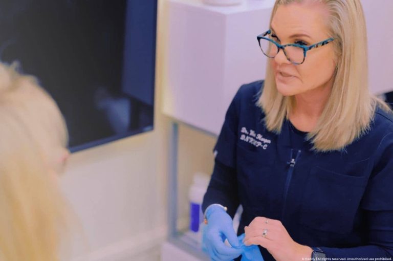 Female aesthetic injector in navy medical scrubs and blue nitrile gloves consulting with a patient in a modern treatment room, representing patient education and informed consent in medical aesthetics clinical practice