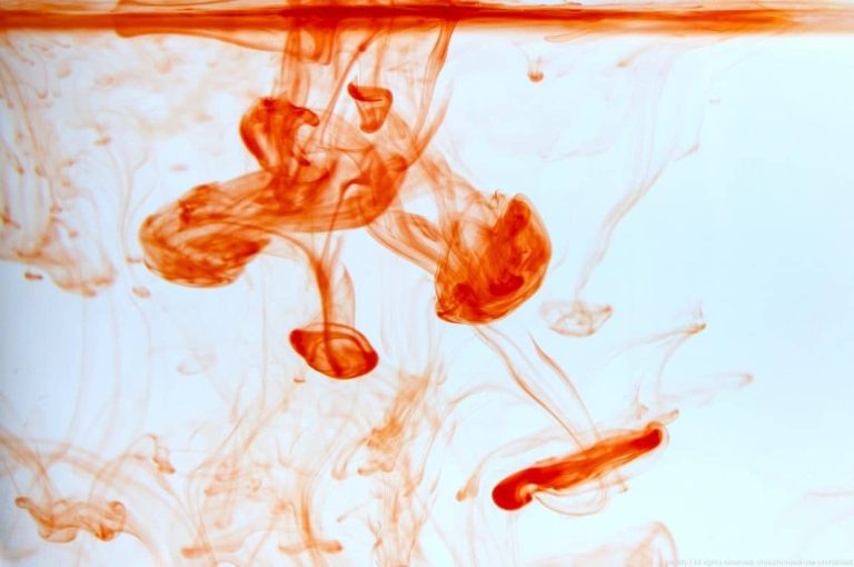 Abstract macro photograph of red-orange ink dispersing in water, representing hyaluronidase enzyme diffusion through hyaluronic acid filler tissue in aesthetic medicine